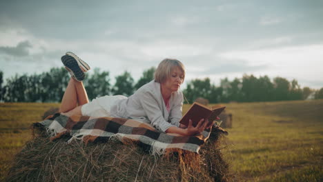 middle-aged woman with leg raised on hay bale with checkered blanket, reading book in open field with strand of hay in her mouth, playing with it in a peaceful, rural setting