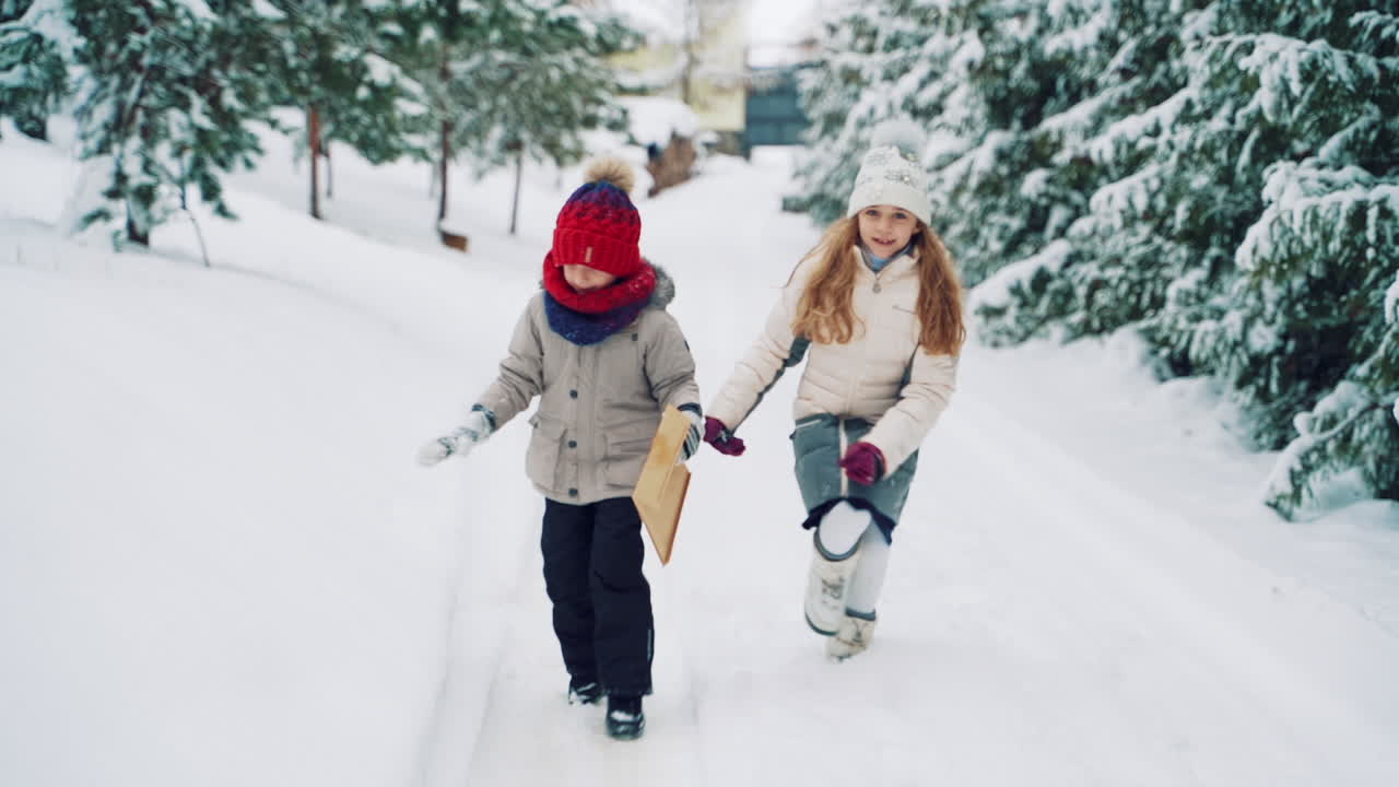 Pretty little girls are walking in the snow on the background of fir trees. Happy children with a big envelope in winter outdoors. Slow motion.
