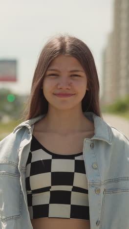 positive teenage girl smiles looking at camera. young female walks in yard of high-rise building on warm summer day closeup on blurred background