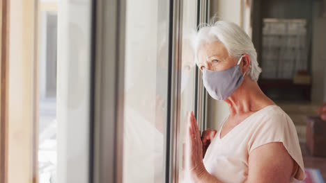 senior caucasian woman wearing face mask looking out of window at home