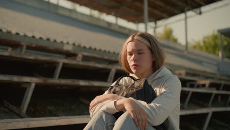 close-up of a young woman sitting alone on empty stadium bleachers, deeply lost in thought, hugging her bag with a reflective expression, with a blurred background