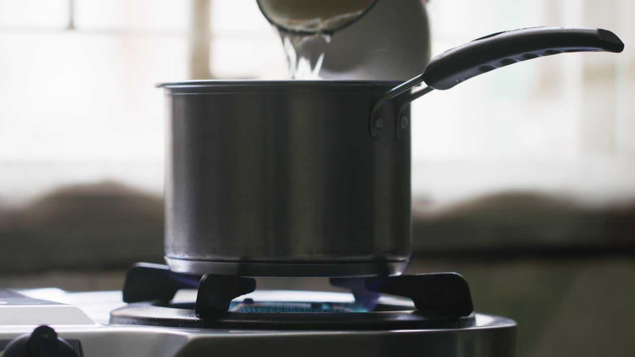 A slow-motion of pouring water into a stainless-steel pot on a lit stovetop against a blurred background