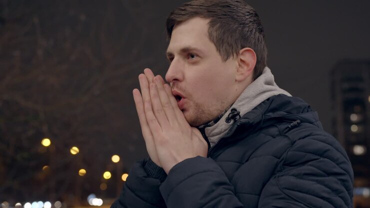 Giovane uomo infreddolito che si riscalda le mani con il fiato, notte fredda d'inverno, scatto a mano libera con spazio per il testo