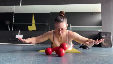 a woman lying face down on the mat in the gym, exercising with her hands in the air, a woman with glasses doing sports