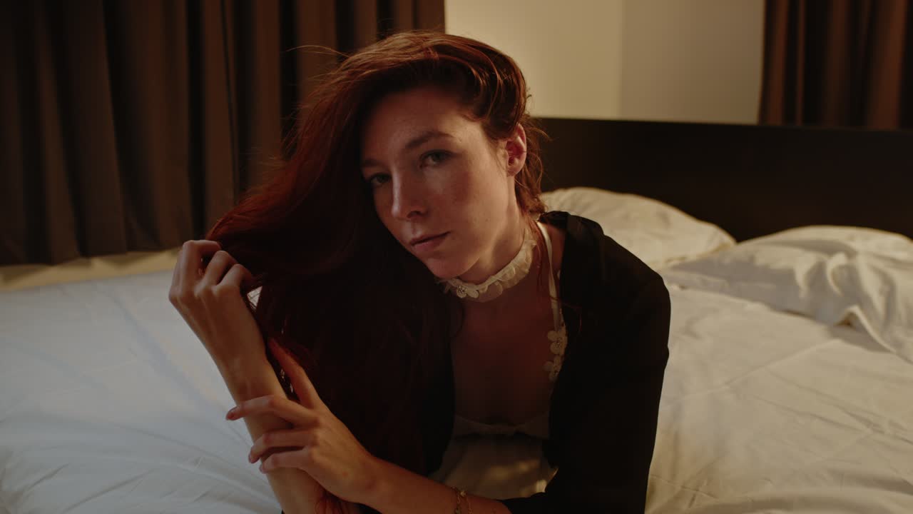 A stunning red haired woman lies in bed and plays with her hair while looking up to the camera