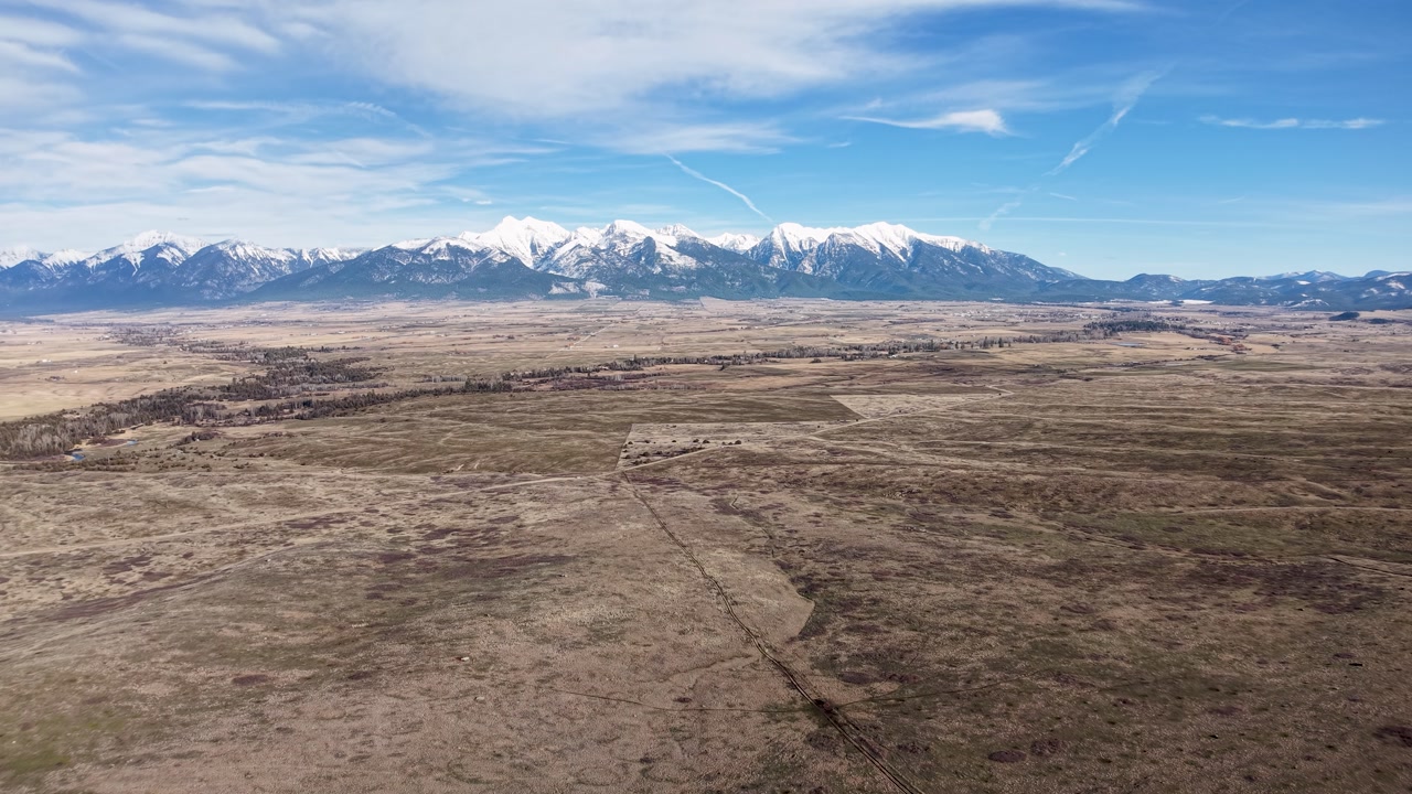 Aerial drone footage flying over a wide, open plain near Whitefish, Montana, reveals a dry prairie with sparse vegetation and snowcapped Rocky Mountains on the horizon under a bright sky.