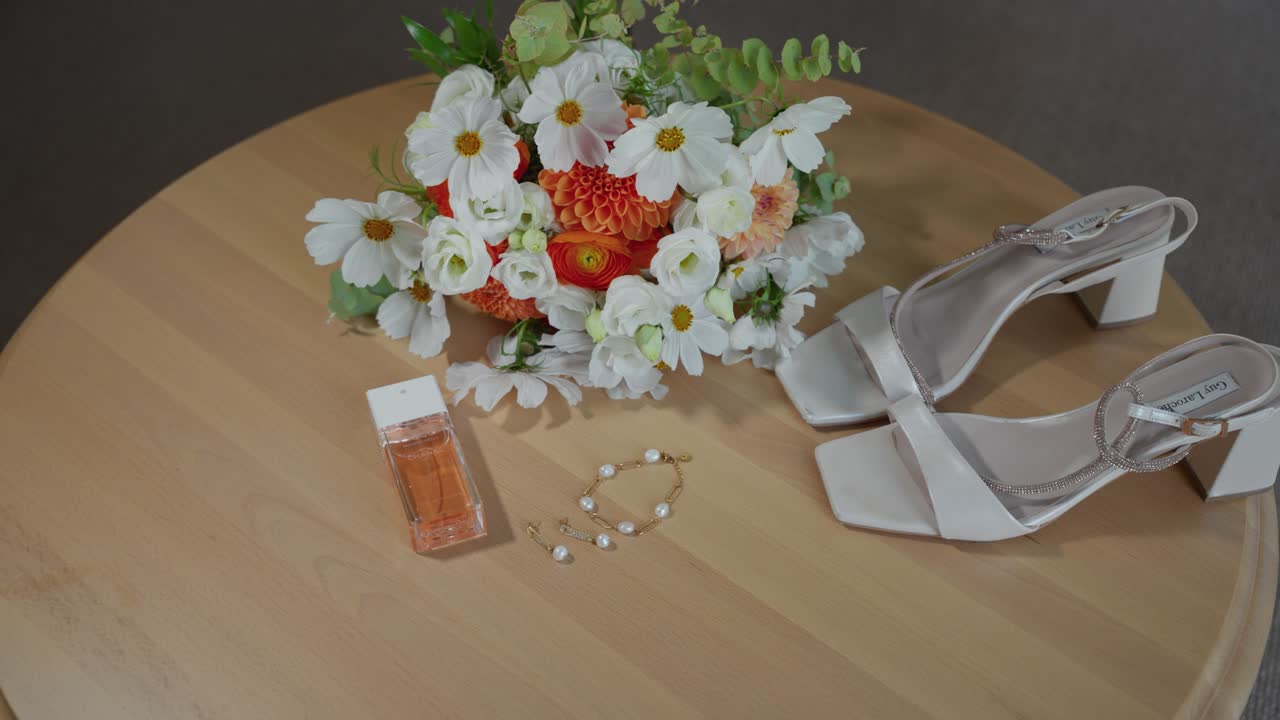 Close-up shot of bridal essentials: white heels, floral bouquet, perfume, and jewelry on a wooden table, showcasing elegance and craftsmanship under soft ambient lighting
