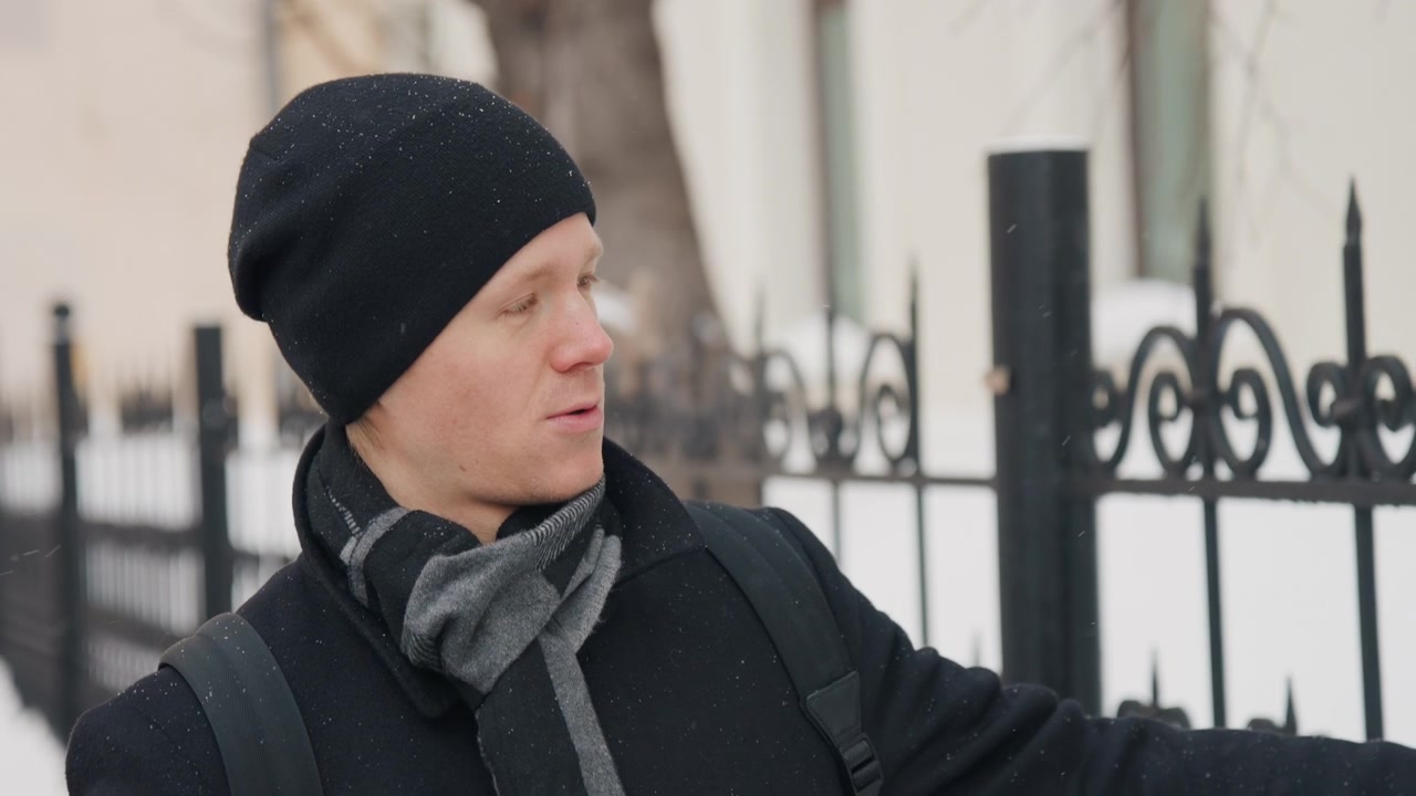 Primo piano di un uomo che indossa abiti invernali, inclusi un cappotto nero, una sciarpa grigia e un berretto, in piedi all'aperto con tempo nevoso. Il soggetto rimane fermo con sottili movimenti della testa contro uno sfondo urbano.
