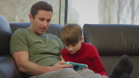 padre con su hijo con pijama sentado en el sofá en casa jugando a un juego de computadora en un dispositivo portátil - filmado en cámara lenta