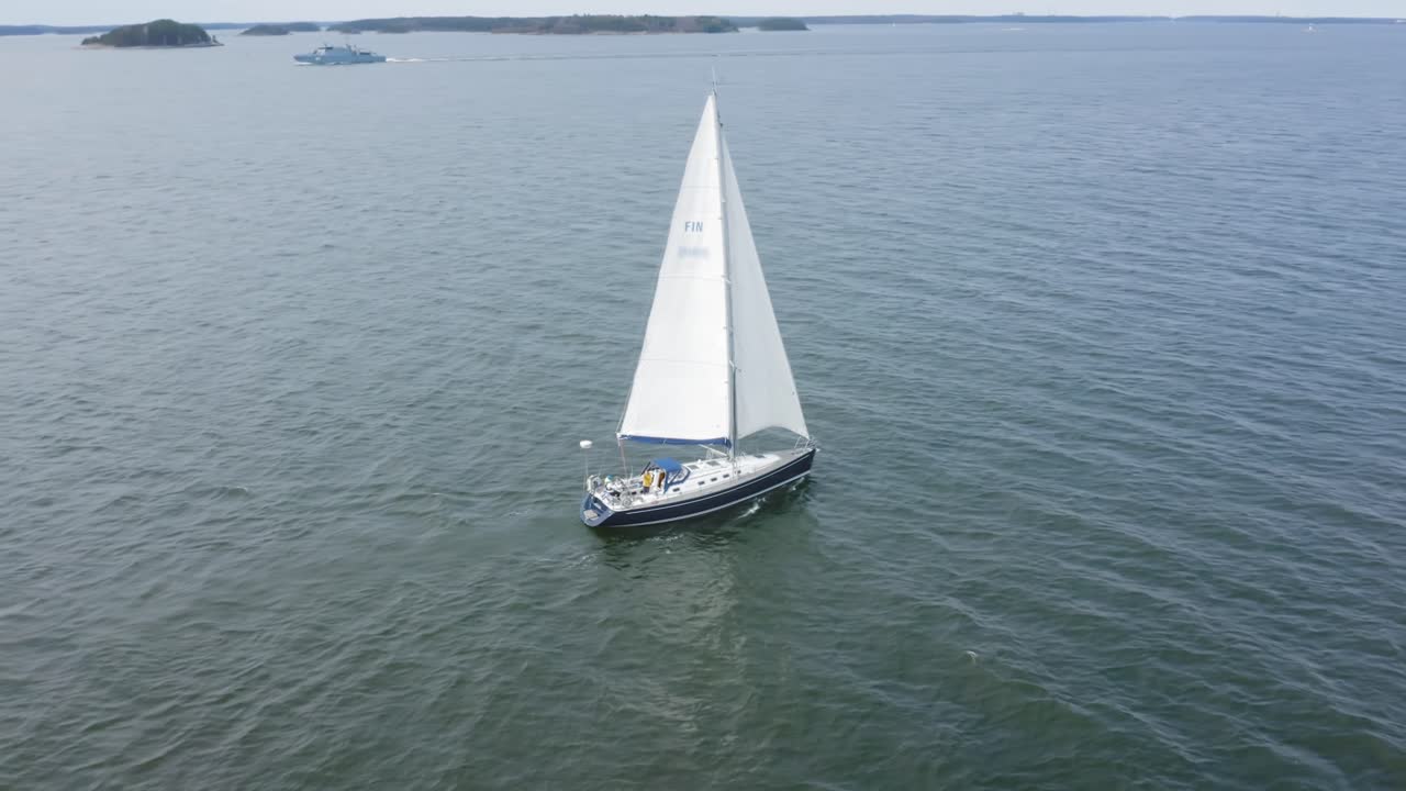 Sailboat navigating with full sails in Finland South West archipelago. Small islands in the background. Aerial drone view circling around. Person waving from the boat at the drone to come closer