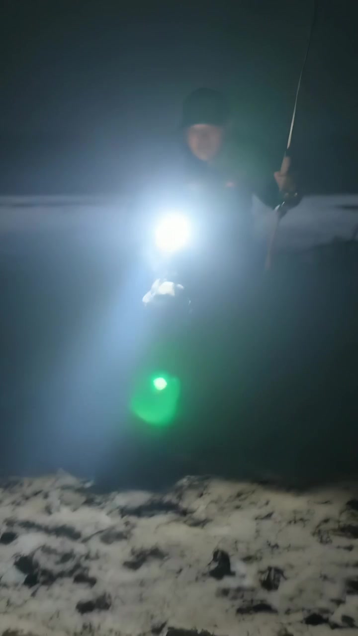 Video vertical: Hombre con abrigo y linterna encendida caminando por la orilla de noche con una caña, buscando dónde pisar.