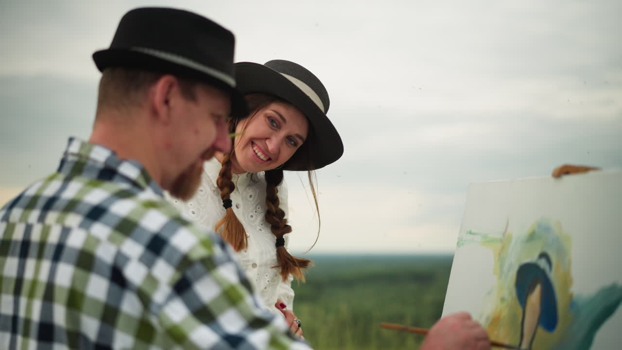 un artista con un sombrero y una camisa a cuadros está profundamente enfocado en pintar al aire libre. una mujer con un vestido blanco lo interrumpe juguetónamente, tocando suavemente su cara con una hoja de hierba