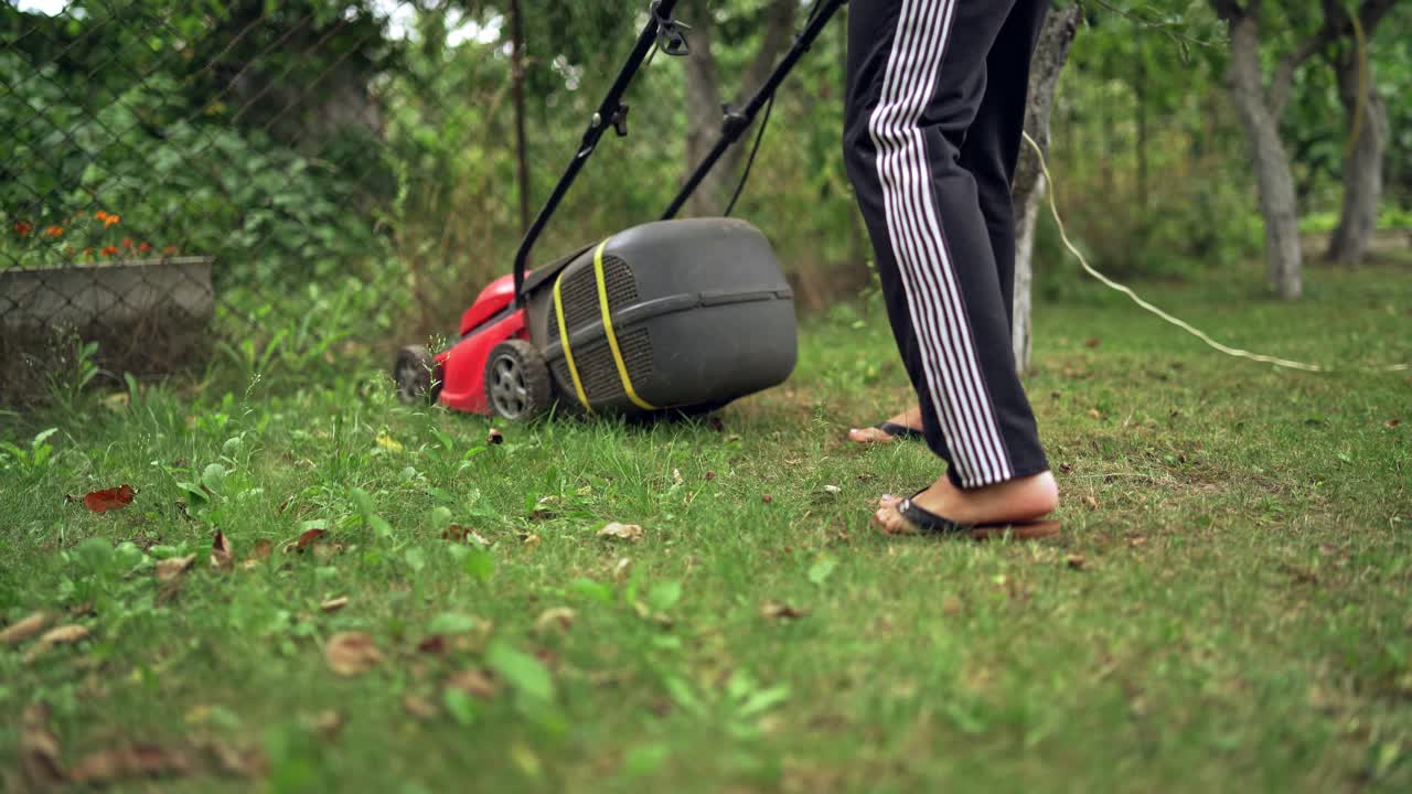 Woman's legs with lawn mover machine outdoors. Lawn mower cutting the grass in a summer day. Gardening activity .