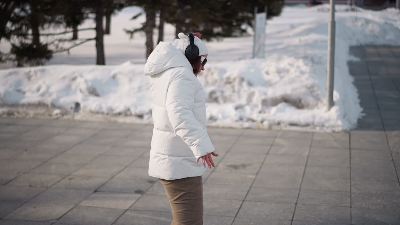 Young girl wearing white winter coat, beanie and headphones demonstrates dance move by turning body and moving hands on tiled plaza with snowbanks and trees under cold daylight in urban park setting
