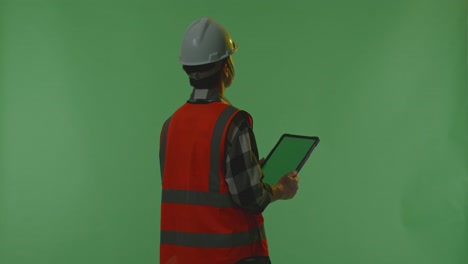 back view of asian male engineer with safety helmet working on a green screen tablet and looking around while standing in the green screen background studio, during sunset or sunrise time