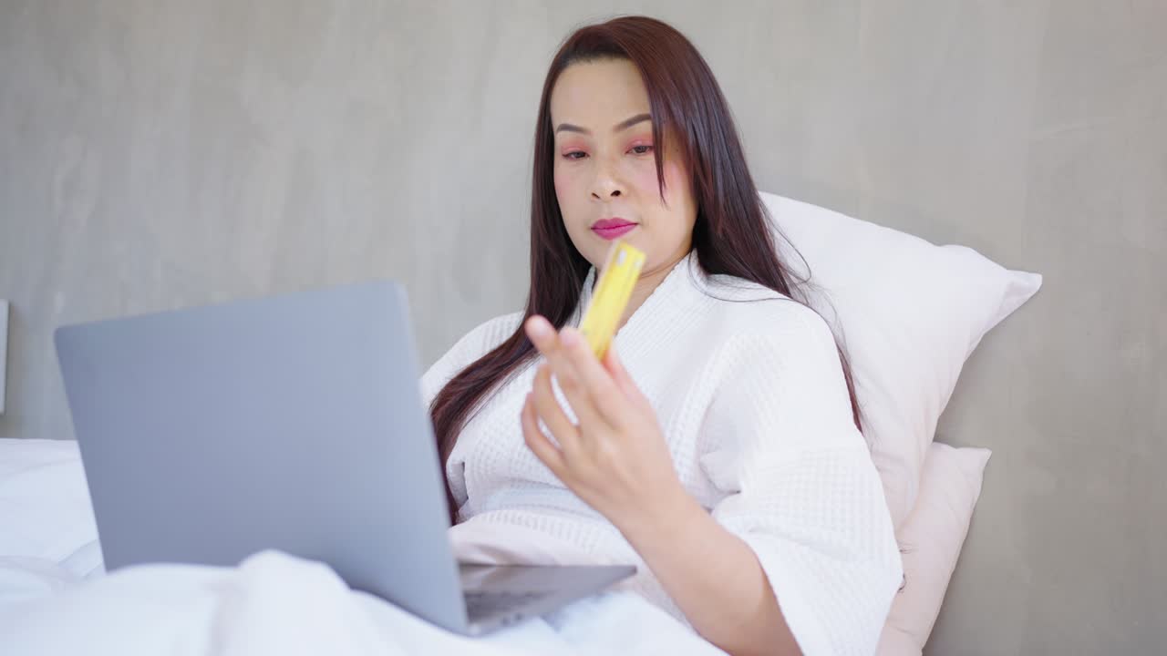아시아 여성이 노트북 컴퓨터와 은행 신용카드로 온라인 쇼핑을 하고, 침실의 편안함 속에 있는 여성