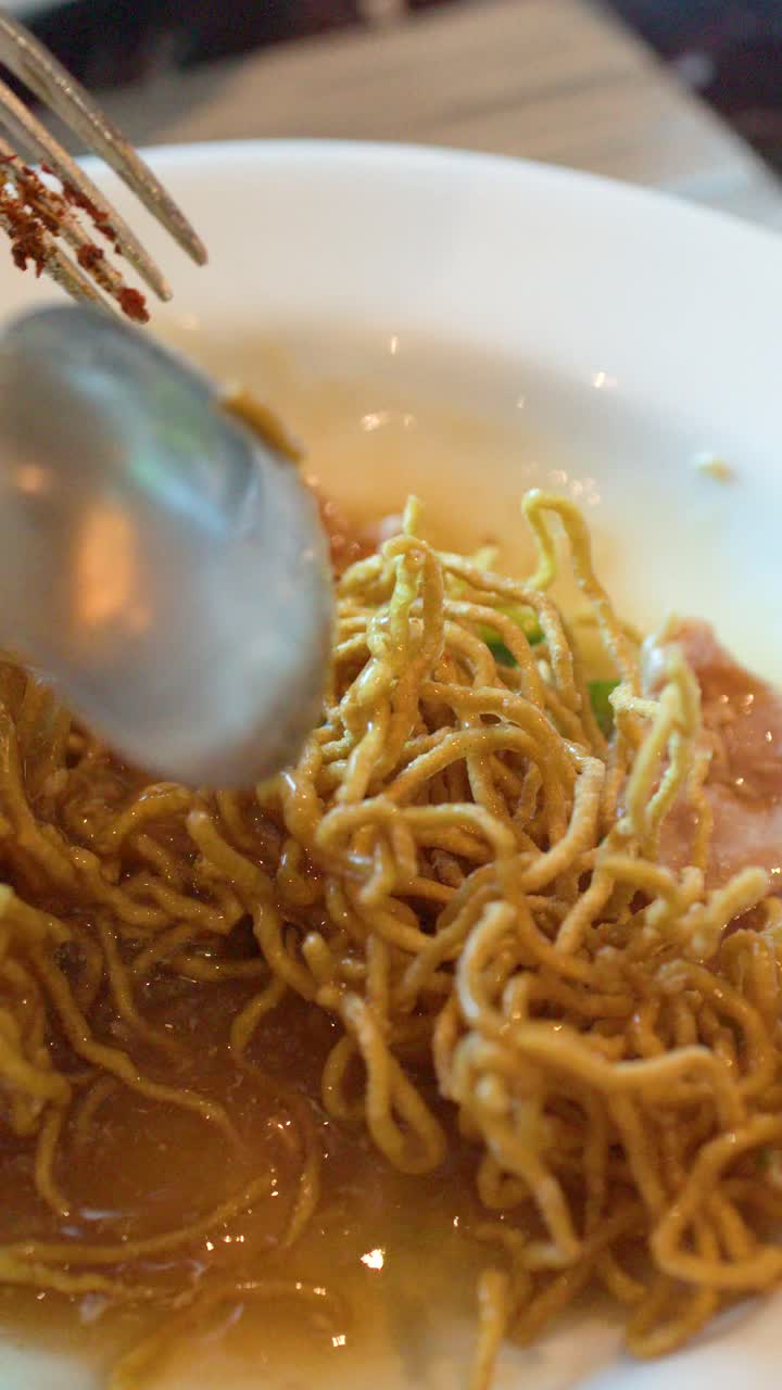 クローズアップショットでは、風味豊かなグレービーソースで豚肉とカリカリの卵麺を混ぜる人が写っている。