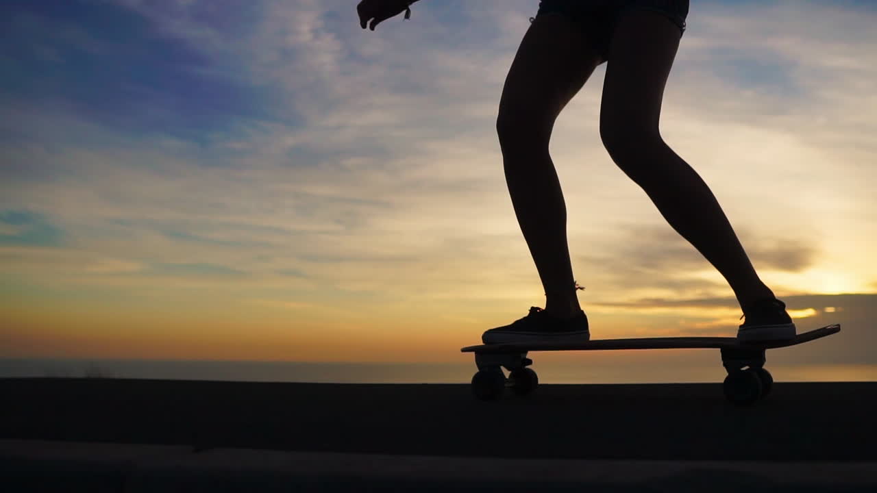 In slow motion, a beautiful and stylish skateboarder cruises on her board in shorts along a mountain road during sunset, showcasing a breathtaking mountain view