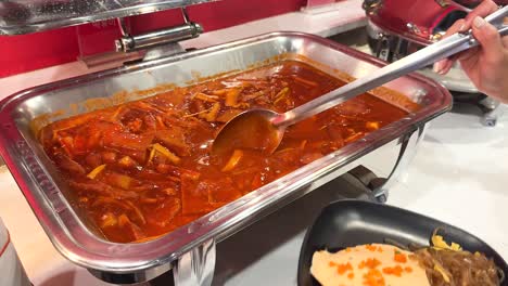 a person serves spicy korean tteokbokki from a buffet tray onto a plate in a well-lit setting