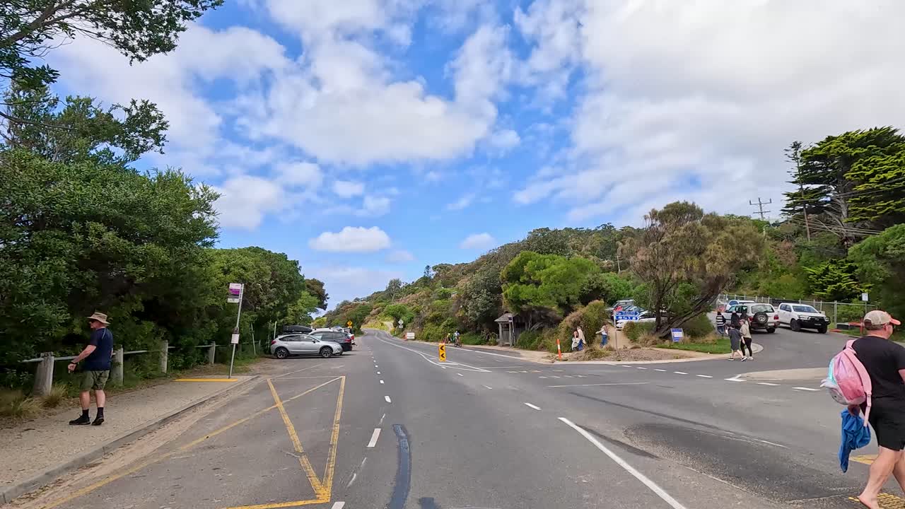 A sunny day on Great Ocean Road with pedestrians, cyclists, and vehicles. Lush greenery and blue skies create a vibrant atmosphere