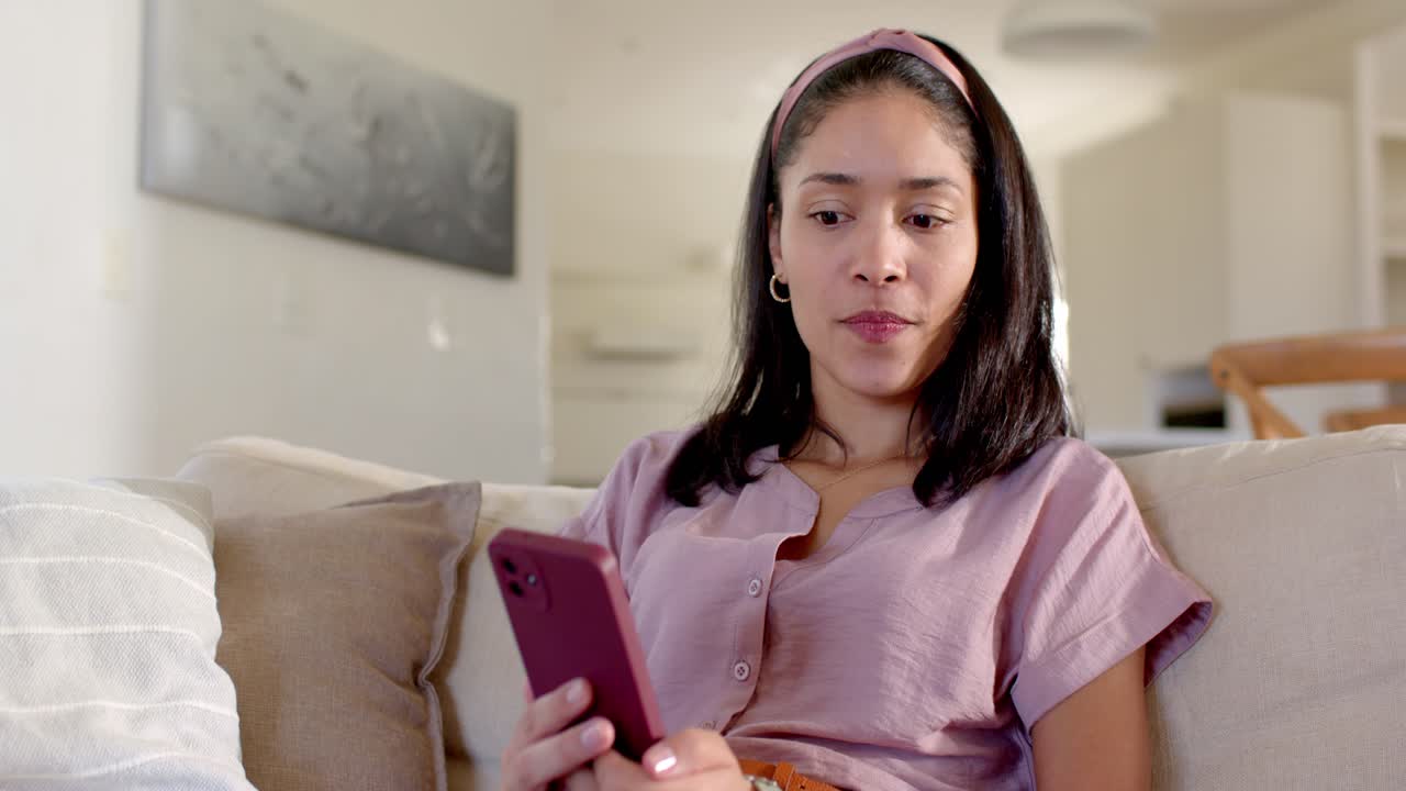 Donna afroamericana legge una notifica, reagisce e guarda a destra, seduta sul divano con il suo smartphone rosa.