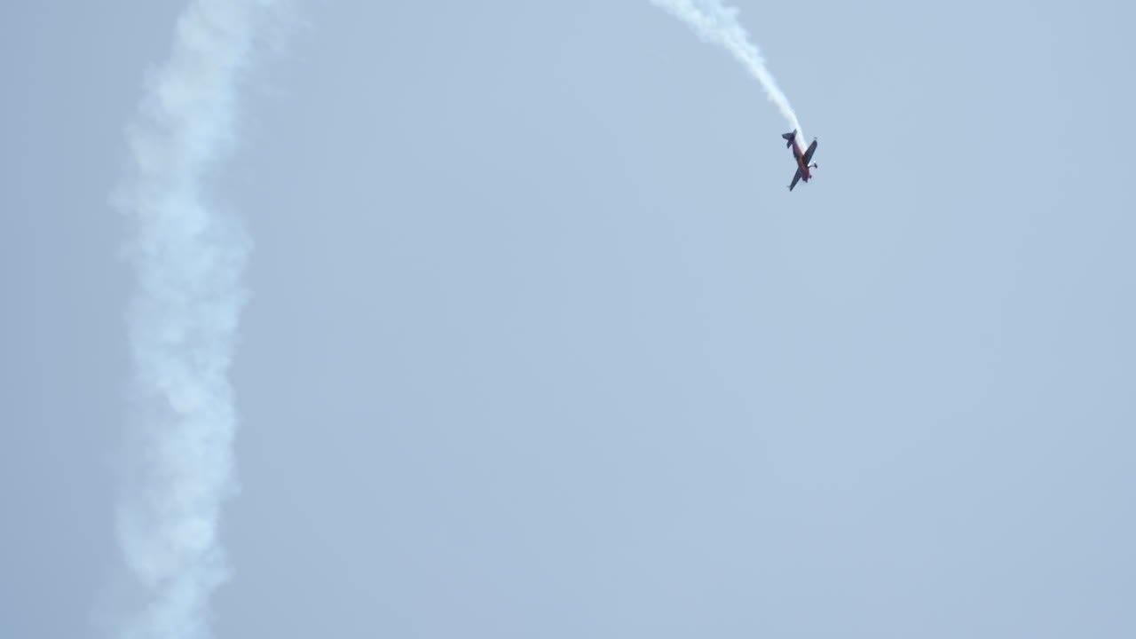 Single red aerobatic airplane climbs vertically with smoke trail during an airshow under clear sky