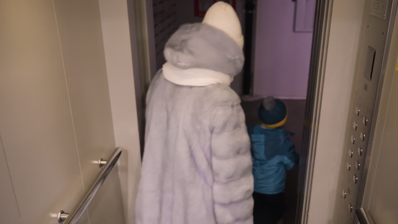 White mom and son exit elevator into dim hallway, mom guides child toward doorway in soft lavender coat and white beanie, small green shovel near threshold, protective walk and quiet evening routine