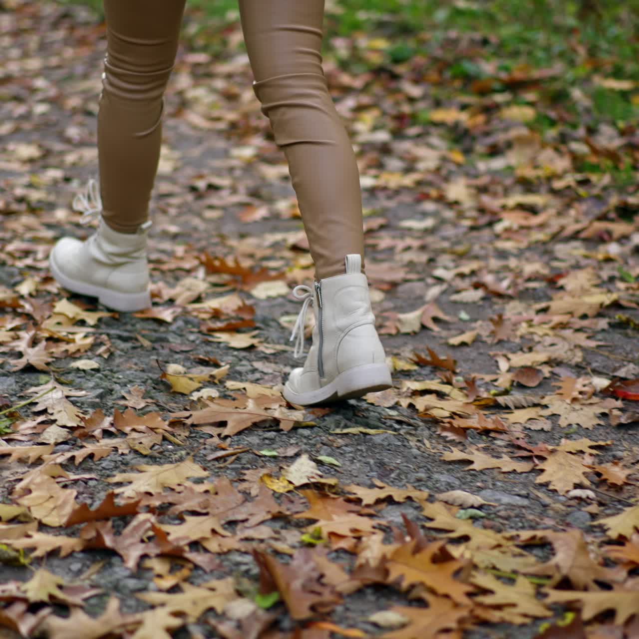 Female in tight beige leggings and white boots walks quickly by the path in the park. Unrecognized lady taking a stroll on autumn day