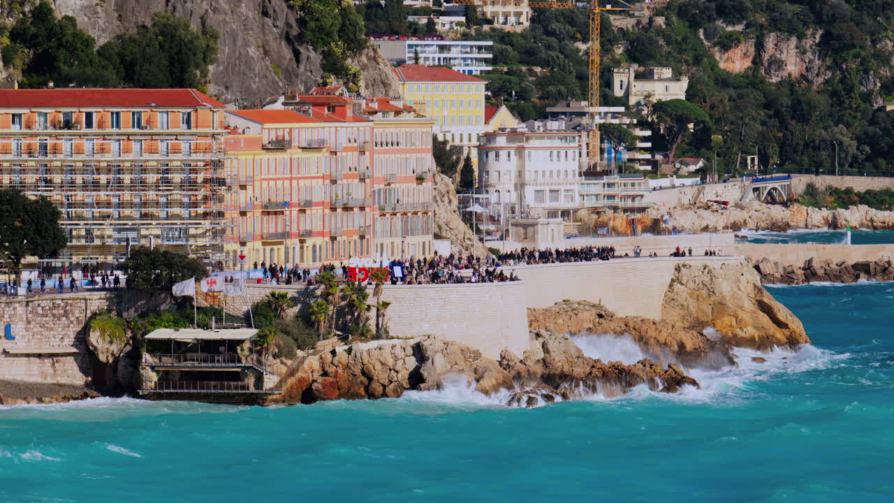 Nice, France - March 17, 2025: View of people walking on the coast of the city with water crashing on the rocks and the buildings on the background