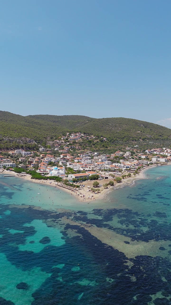 Ripresa panoramica verticale con drone del villaggio e della spiaggia di Skala, Isola di Agistri, Grecia. Ombrelloni da spiaggia e bagnanti visibili. Giornata estiva limpida e soleggiata.