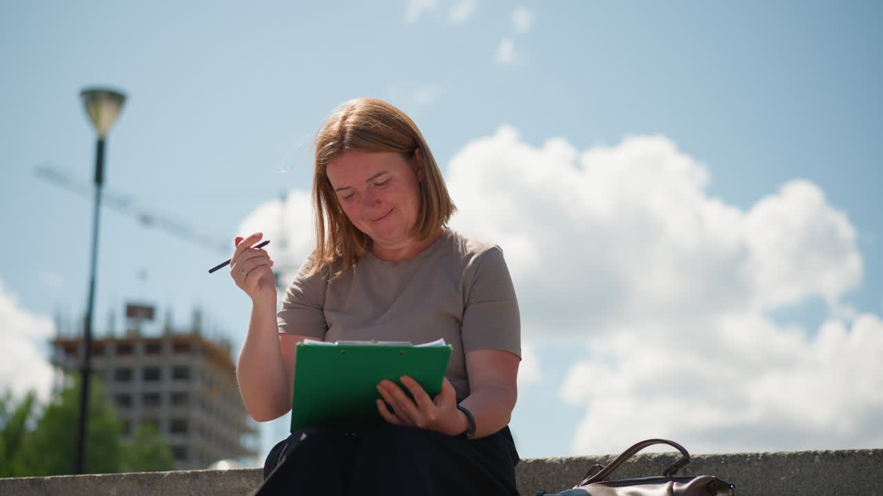Office assistant sitting alone on stone steps writing on clipboard under warm sunlight, wearing beige top and black skirt, modern building in background, showing concentration