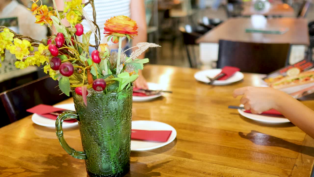 Hands arrange plates and napkins on a wooden table in a brightly lit restaurant with floral centerpiece