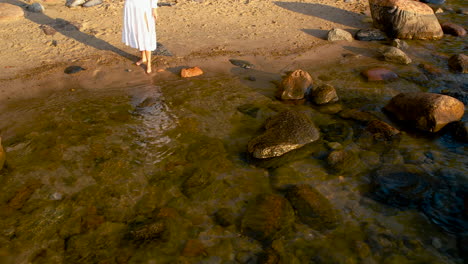 an unrecognizable woman in a white dress standing by rocky seashore with stones in sea water at sunrise