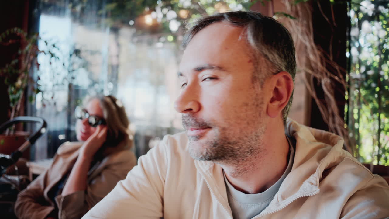 Primo piano di un uomo che sorseggia un caffè alla luce naturale del sole, vicino a una donna che indossa occhiali da sole in un accogliente bar all'aperto