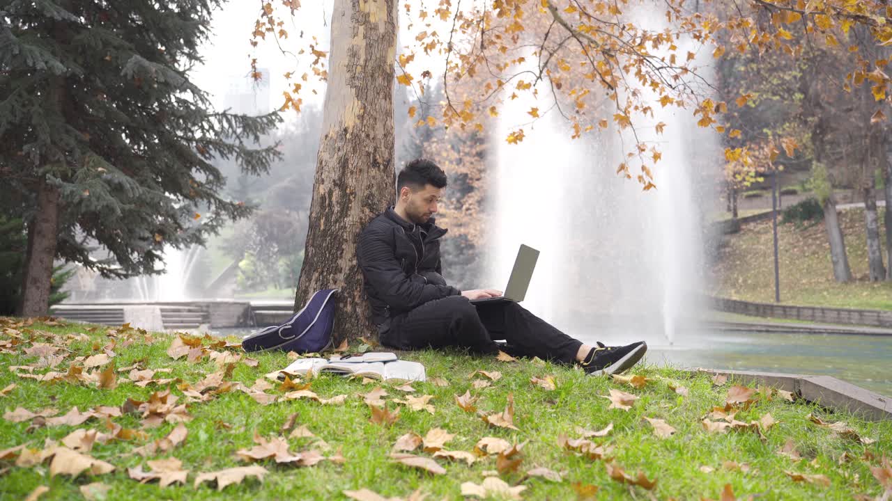 도시 공원에서 잔디에 앉아 노트북을 사용하는 남성 학생.