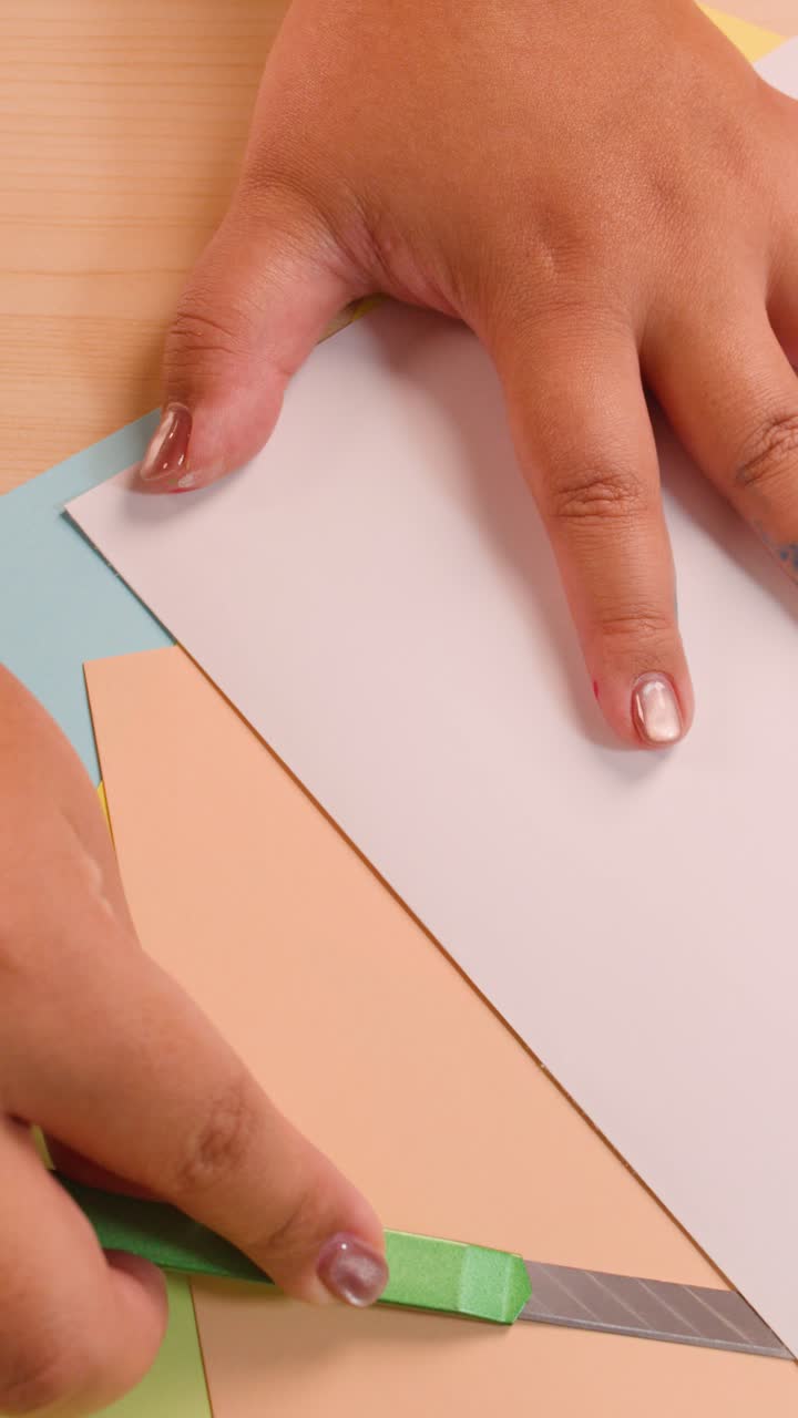 Mani che tagliano e piegano carta colorata su una scrivania di legno, vista dall'alto, con illuminazione naturale soffusa.