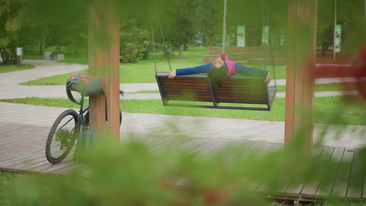 Kvinde slumrer i trægyngepavillon, cykel læner op ad stolpe, rygsæk ved siden af hjul, blødt sommerlys filtrerer gennem træerne, langsom vuggende bevægelse, tom sti i baggrunden, fredfyldt ensomhed
