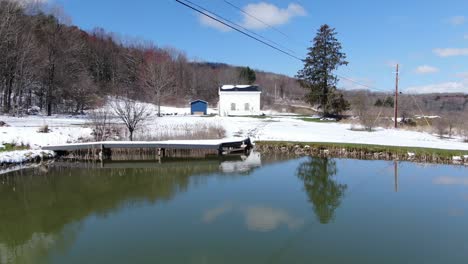 aerial drone footage flying low over a pond towards an old, white house on the edge of a forest covered in snow