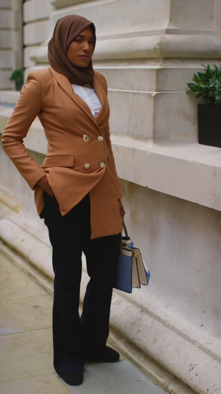 вертикальный полноформатный портрет мусульманской бизнесменки в хиджабе и современном деловом костюме, стоящей за пределами городских офисных зданий 3