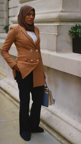 vertical video full length portrait of muslim businesswoman wearing hijab and modern business suit standing outside city office buildings 3