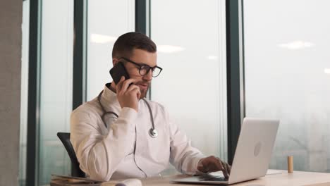 Attractive-young-doctor-sitting-behind-a-laptop-is-talking-on-the-phone-with-a-client