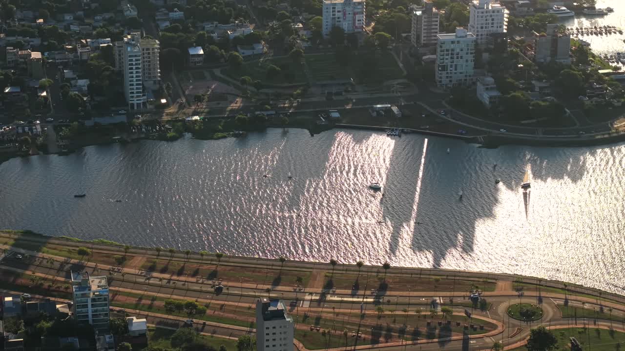 Bahía El Brete in Posadas, Misiones province, Argentina, seen in a slow drone pan with sunlight reflecting on the parana river bay, tall buildings casting long shadows and boats visible near the shore
