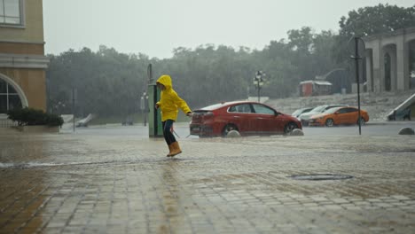 Happy-teenage-girl-in-a-yellow-jacket-and-orange-rubber-boots-runs-through-streams-of-water-and-puddles-during-heavy-rain-outdoors