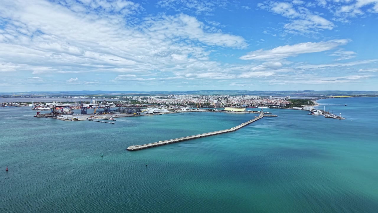 Vista costiera di un porto trafficato vicino a una città con navi, banchine, acqua e nuvole nel cielo, che mostra attività urbana durante il giorno.