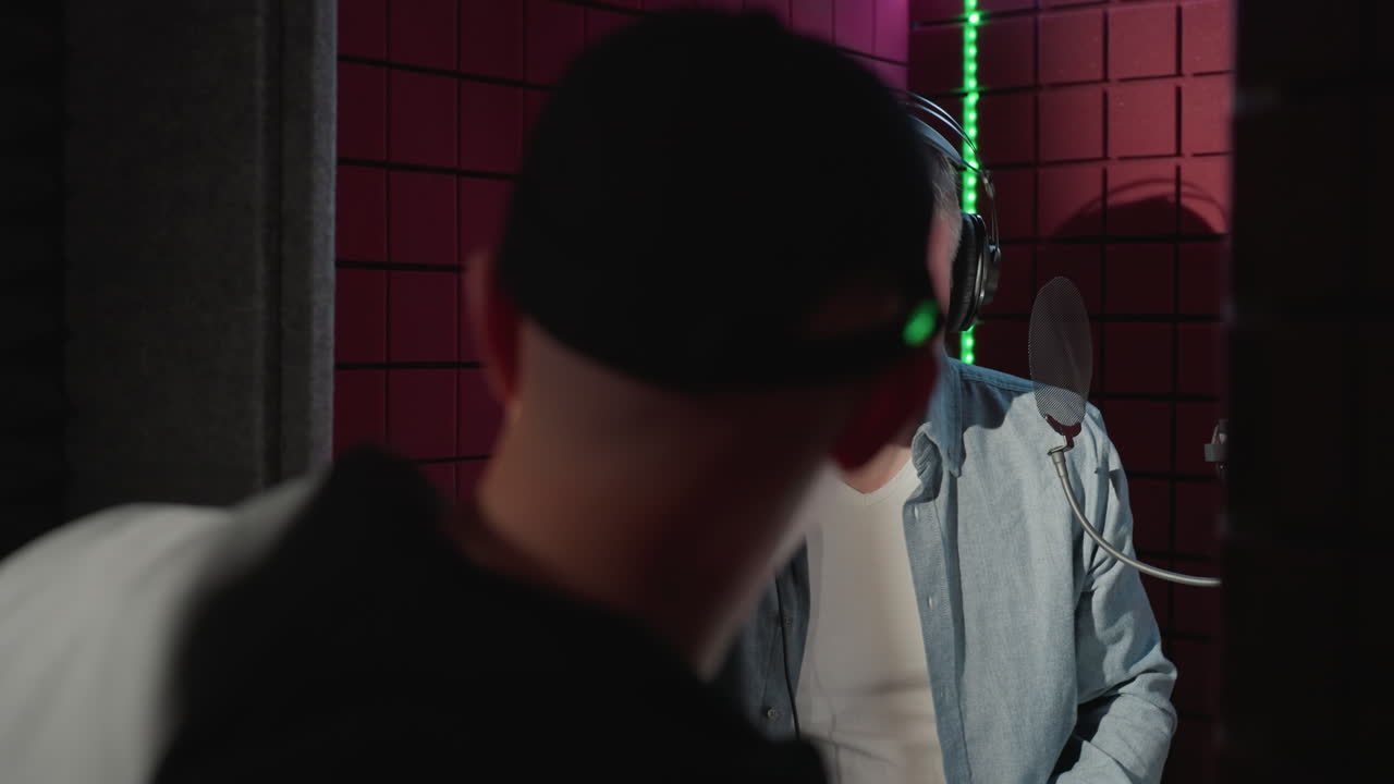 en un estudio a prueba de sonido rojo, un hombre con una camisa negra ayuda a otro con una camiseta azul de manga larga, colocándole auriculares mientras canta alegremente, preparándose para una sesión de grabación