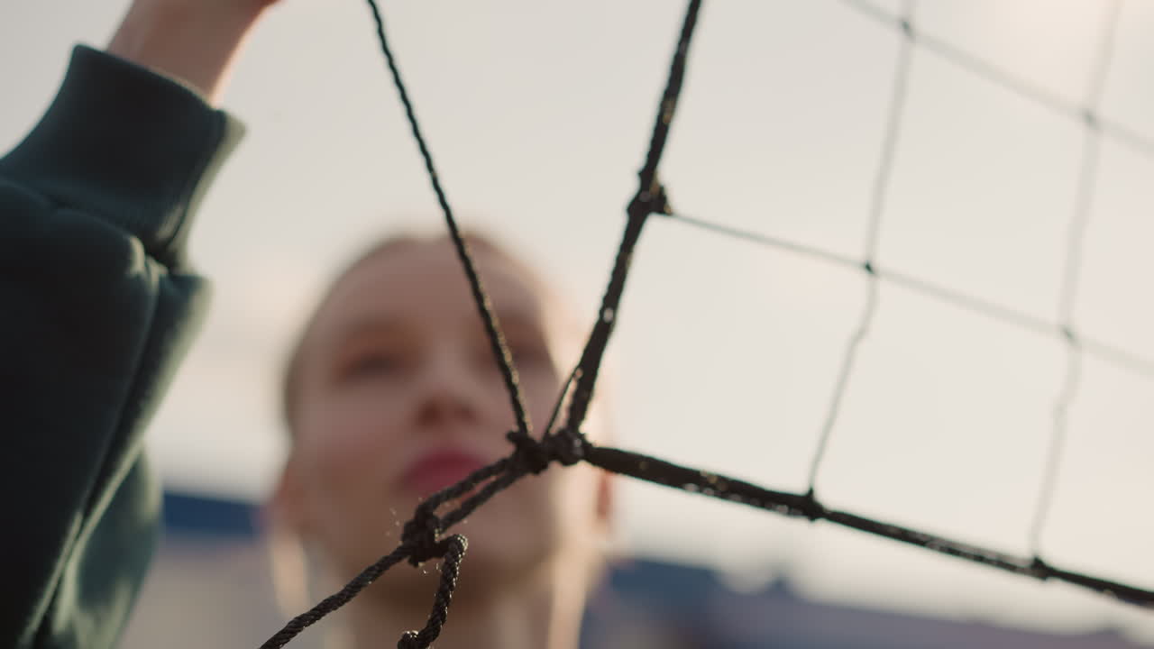 Close-up of volleyball net held by someone in green sweater, checking it with hazy sky in background, focusing on net and hands, face slightly blurred