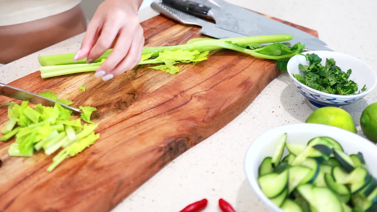 Hands skillfully chop celery on a wooden board, surrounded by fresh ingredients in a well-lit kitchen