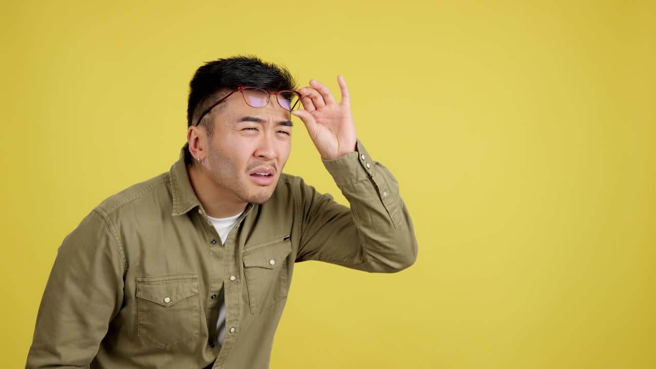 Young Man with Glasses Expresses Surprise, Listens, and Adjusts Glasses on Yellow Background