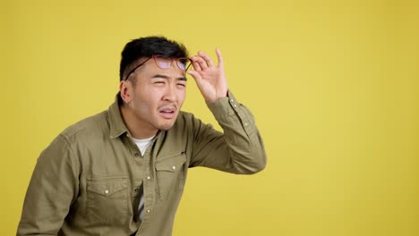 young man with glasses expresses surprise, listens, and adjusts glasses on yellow background