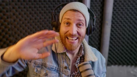 camera point of view - young man with headphones records message in sound studio. happy enthusiastic excited hipster podcast or radio host talking to camera waving hello and speaking live to webcam